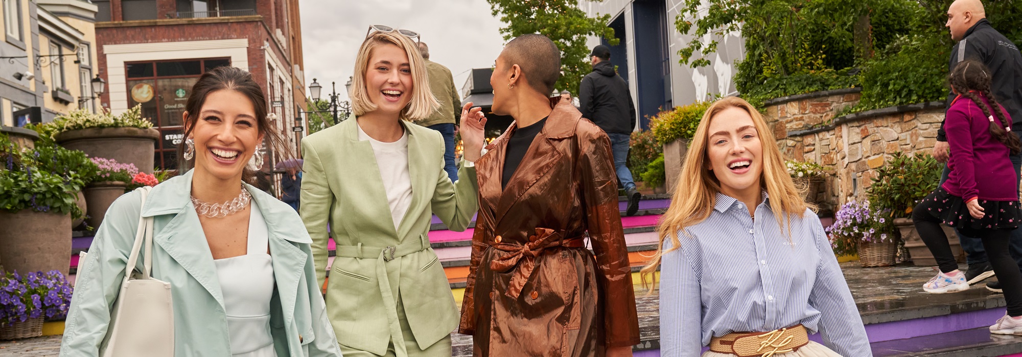 Vier vrouwen lopen lachend de kleurrijke trappen af bij Designer Outlet Roermond, gekleed in modieuze outfits, met winkels en bezoekers op de achtergrond.