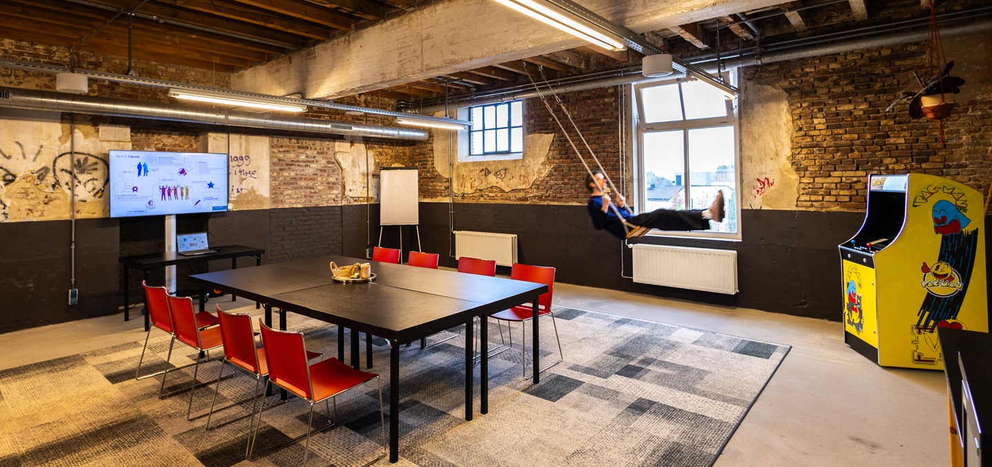 Industriële ruimte bij ECI in Roermond met bakstenen muren, een grote vergadertafel met rode stoelen, een schommel en een gele Pac-Man-arcadekast.