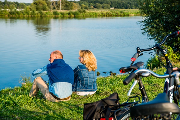 Vrouw en man zitten aan de oevers van de Maas, uit te rusten tijdens hun fietstocht door de gemeente Leudal