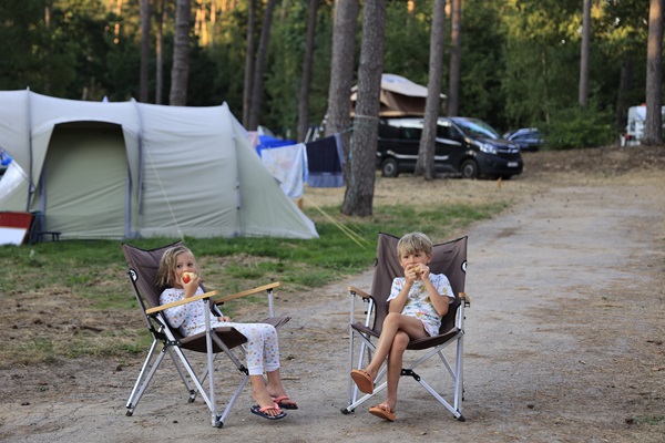 Twee kinderen zitten in kampeerstoelen en eten een appel op een camping bij Huttopia in de gemeente Roerdalen, met tenten en bomen op de achtergrond.