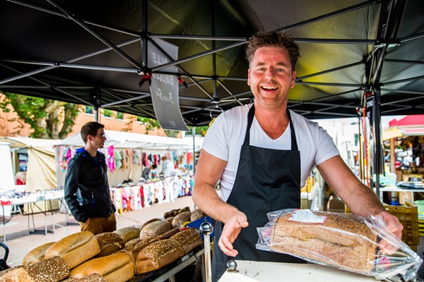 Lächelnder Marktverkäufer auf einem Wochenmarkt in Limburg, der frisch gebackenes Brot unter einem Marktstand anbietet.