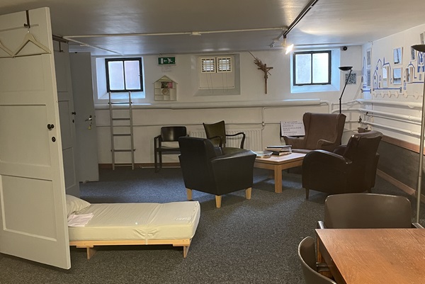 Kleine, knusse verblijfsruimte met fauteuils, een tafel en een eenvoudig bed, ingericht als pelgrimsopvang in Roermond.