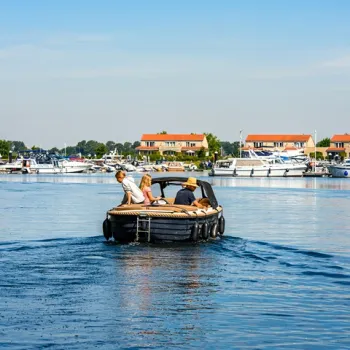 Gezin in een sloep op het water, met jachthaven en woningen op de achtergrond onder een heldere lucht.