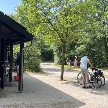 Mensen met fietsen bij Natuurkampeerterrein Wega, omringd door bomen en een picknickplek.